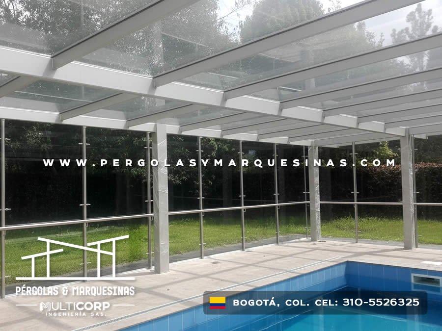 Pérgola de vidrio con columnas blancas y cerramiento lateral junto a piscina en jardín residencial en Bogotá, Colombia. Multicorp Ingeniería SAS.