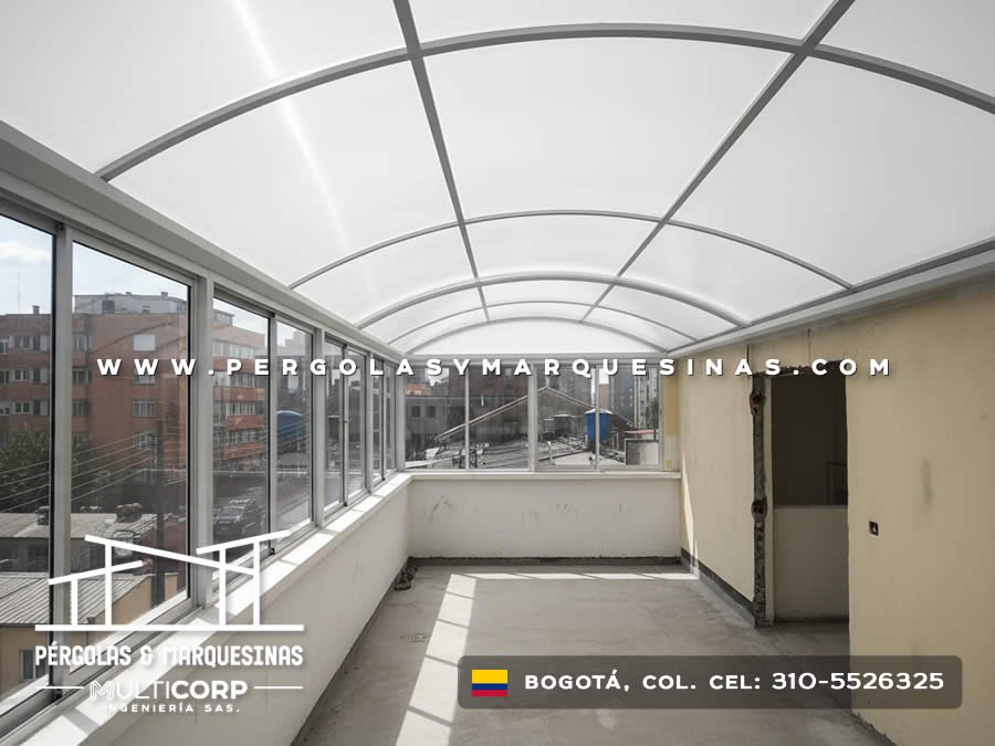 Cubierta arqueada de policarbonato con estructura gris sobre balcon con ventanales y vista a edificios en Bogotá, Colombia. Multicorp Ingeniería SAS. Cubierta arqueada de policarbonato con estructura gris sobre balcon con ventanales y vista a edificios en Bogotá, Colombia. Multicorp Ingeniería SAS.