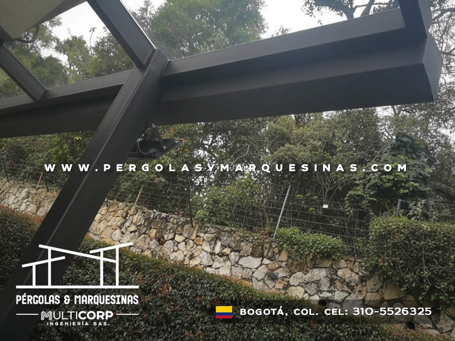 Pergola acristalada metalica negra techo vidrio sobre jardin con muro piedra natural y vegetacion frondosa en Bogotá, Colombia. Multicorp Ingeniería SAS. Pergola acristalada metalica negra techo vidrio sobre jardin con muro piedra natural y vegetacion frondosa en Bogotá, Colombia. Multicorp Ingeniería SAS.