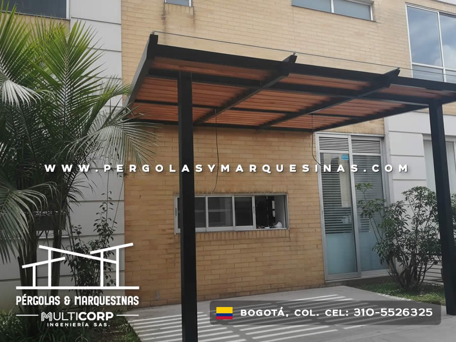 Pergola metalica negra con lamas de madera sobre entrada edificio ladrillo amarillo con palmera en Bogotá, Colombia. Multicorp Ingeniería SAS. Pergola metalica negra con lamas de madera sobre entrada edificio ladrillo amarillo con palmera en Bogotá, Colombia. Multicorp Ingeniería SAS.