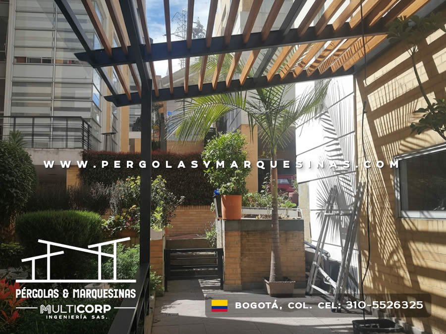 Pergola metalica negra con lamas de madera sobre jardin urbano con palmera macetas y edificios al fondo en Bogotá, Colombia. Multicorp Ingeniería SAS. Pergola metalica negra con lamas de madera sobre jardin urbano con palmera macetas y edificios al fondo en Bogotá, Colombia. Multicorp Ingeniería SAS.