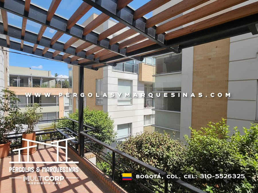 Pergola metalica negra con lamas de madera sobre terraza balcon con baranda edificios urbanos y vegetacion en Bogotá, Colombia. Multicorp Ingeniería SAS. Pergola metalica negra con lamas de madera sobre terraza balcon con baranda edificios urbanos y vegetacion en Bogotá, Colombia. Multicorp Ingeniería SAS.