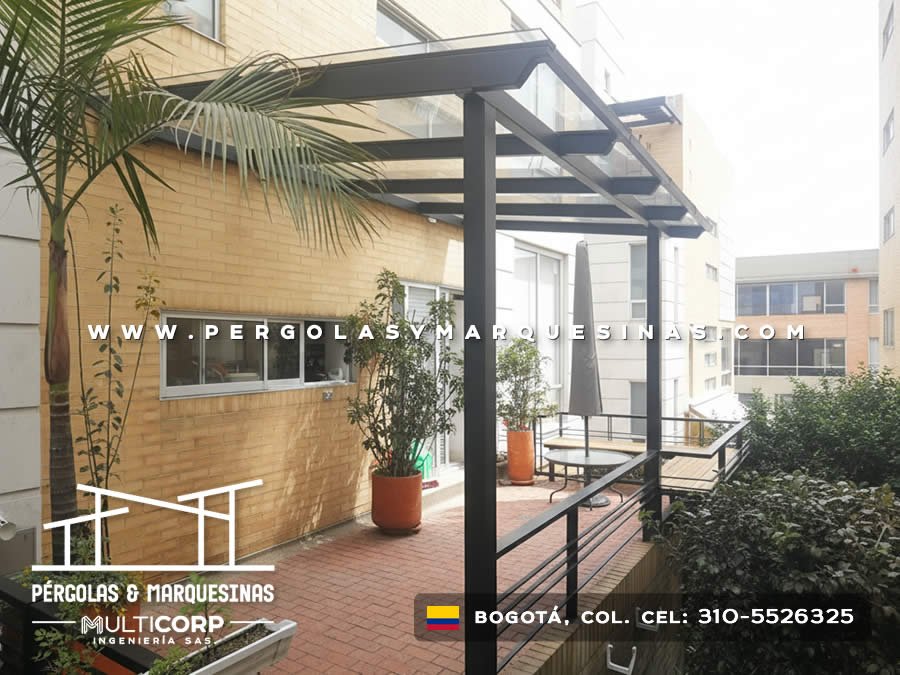 Pergola metalica negra techo policarbonato sobre jardin terraza edificio ladrillo amarillo con palmera macetas y baranda en Bogotá, Colombia. Multicorp Ingeniería SAS. Pergola metalica negra techo policarbonato sobre jardin terraza edificio ladrillo amarillo con palmera macetas y baranda en Bogotá, Colombia. Multicorp Ingeniería SAS.