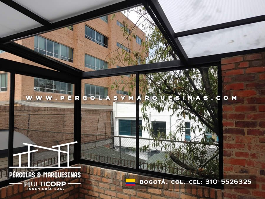 Pergola metalica negra con techo translucido y paneles de vidrio sobre patio residencial con arbol y edificios en Bogotá, Colombia. Multicorp Ingeniería SAS.