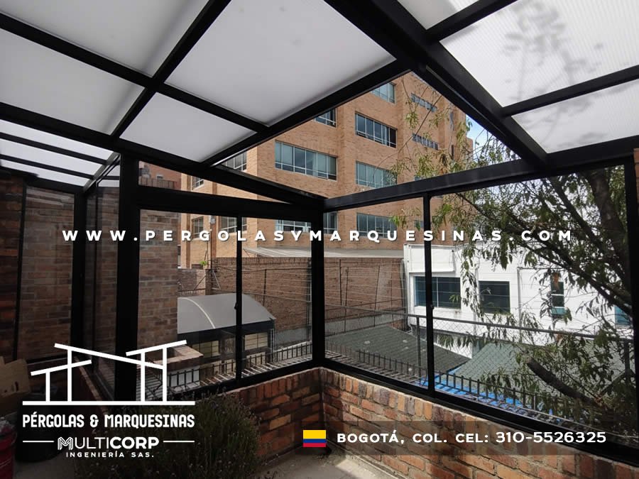 Pergola metalica negra con techo de policarbonato y ventanas de vidrio sobre balcon residencial con vegetacion en Bogotá, Colombia. Multicorp Ingeniería SAS.