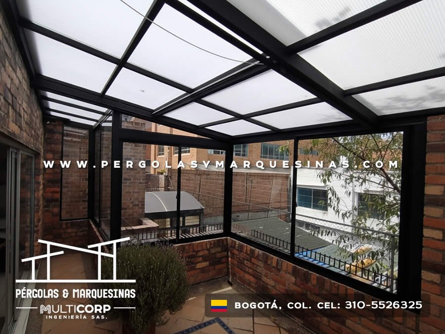 Pergola negra con techo translucido y ventanas de vidrio sobre balcon con jardineras y edificios circundantes en Bogotá, Colombia. Multicorp Ingeniería SAS.