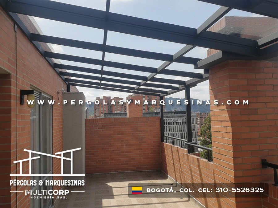 Pergola con techo de vidrio y estructura metalica oscura en terraza de atico con muro de ladrillo y vista urbana en Bogotá, Colombia. Multicorp Ingeniería SAS.