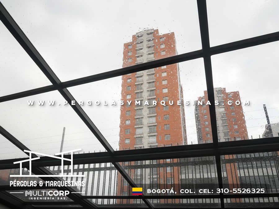 Pergola con estructura metalica negra y cubierta de vidrio bajo lluvia con edificios altos al fondo en Bogotá, Colombia. Multicorp Ingeniería SAS.