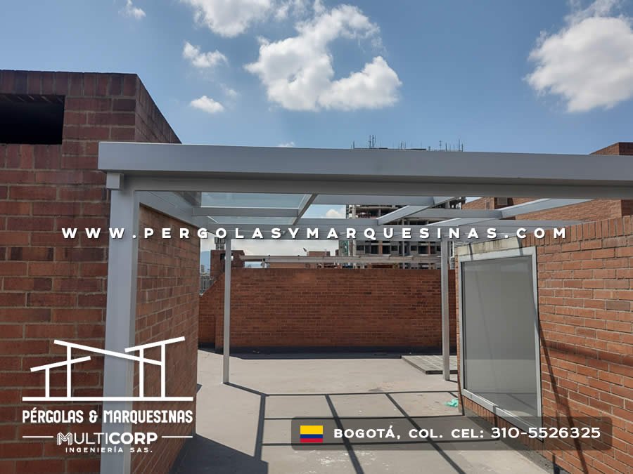 Pergola con estructura gris y cubierta de vidrio en azotea entre muros de ladrillo con vista urbana en Bogotá, Colombia. Multicorp Ingeniería SAS.