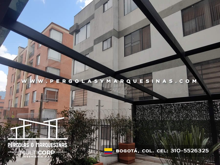 Pergola con estructura negra y vidrio sobre area ajardinada con vegetacion y edificios residenciales en Bogotá, Colombia. Multicorp Ingeniería SAS.