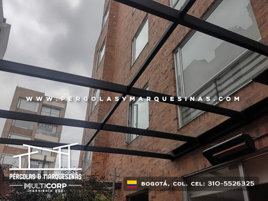 Pergola con estructura negra y vidrio frente a fachada de ladrillo con ventanas y balcones en Bogotá, Colombia. Multicorp Ingeniería SAS.